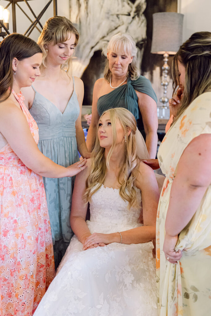 Bride sitting in a chair with all the bridesmaids and her mother around her praying 