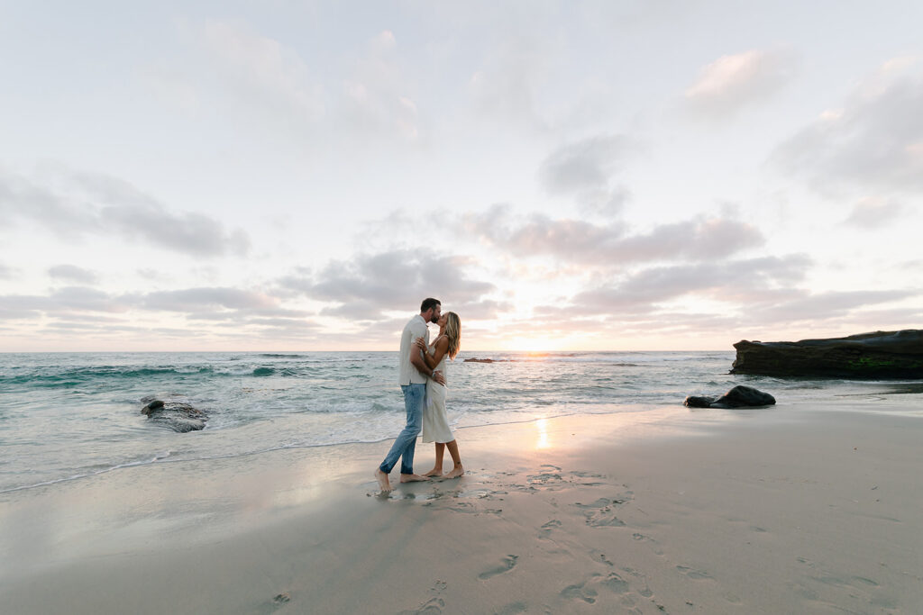 Couple kissing at WindeandSea Beach La Jolla with the sun going  behind the horizon line and pastel tone colors in the sky